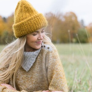 May include: A woman wearing a yellow knitted beanie and a yellow and grey sweater sits in a field of tall grass.