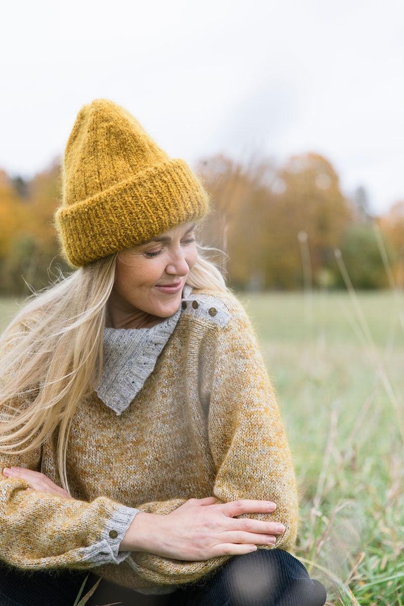 May include: A woman wearing a yellow knitted beanie and a yellow and grey sweater sits in a field of tall grass.