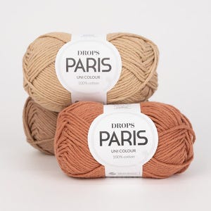 K&ouml;nnte beinhalten: Drei Kn&auml;uel Drops Paris Uni Colour 100% Baumwollgarn in Beige- und Terrakotta-T&ouml;nen. Jedes Kn&auml;uel hat ein wei&szlig;es Etikett mit dem Markennamen und Produktdetails. Das Garn ist fest gewickelt und wirkt weich.