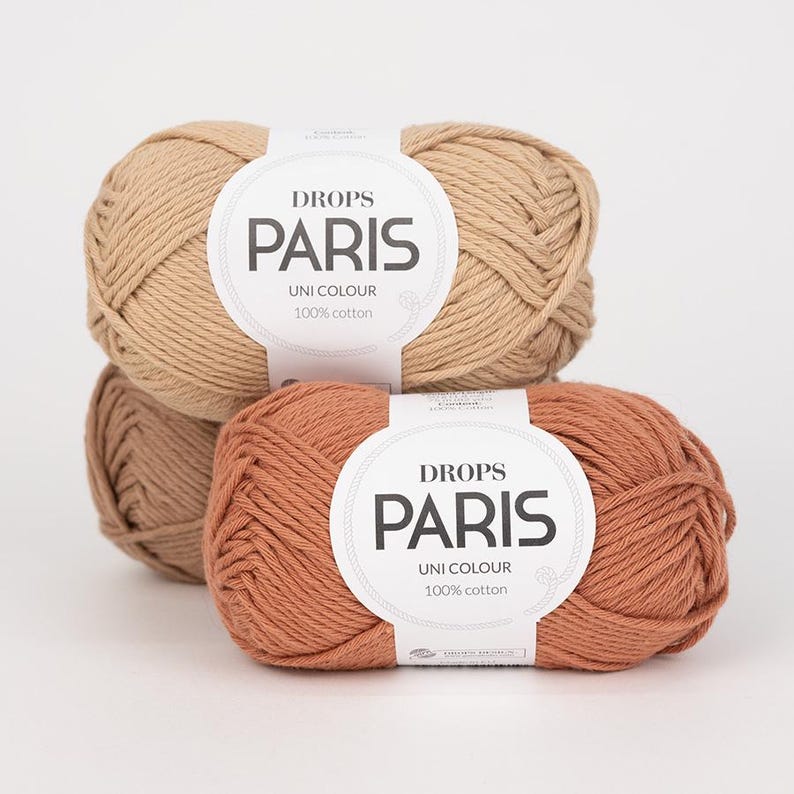 K&ouml;nnte beinhalten: Drei Kn&auml;uel Drops Paris Uni Colour 100% Baumwollgarn in Beige- und Terrakotta-T&ouml;nen. Jedes Kn&auml;uel hat ein wei&szlig;es Etikett mit dem Markennamen und Produktdetails. Das Garn ist fest gewickelt und wirkt weich.