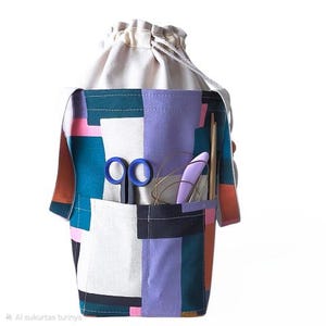 Puede incluir: Una colorida bolsa de manualidades con cierre de cordón. La bolsa presenta un diseño de patchwork en verde azulado, rosa, morado y blanco. Se ven tijeras azules, una herramienta morada y otros suministros de manualidades.