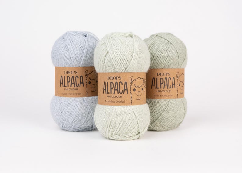 May include: Three skeins of yarn in light blue, pale mint green, and sage green. Each skein features a brown paper label with the text DROPS ALPACA UNI COLOUR and a line drawing of an alpaca face, wrapped around the center of the yarn ball.