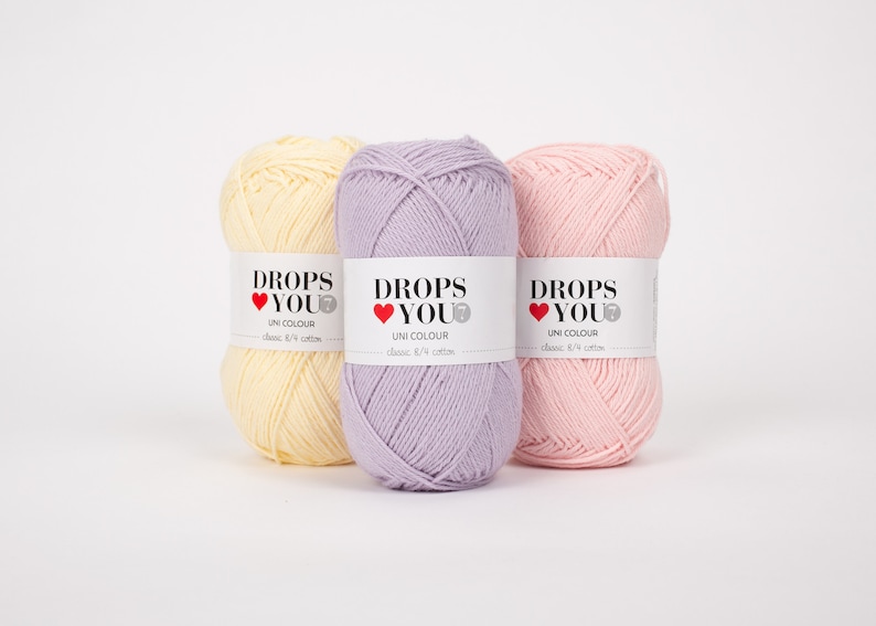 Puede incluir: Tres ovillos de hilo Drops You Uni Colour en tonos pastel: amarillo p&aacute;lido, lavanda y rosa claro. Cada ovillo tiene una etiqueta blanca con el texto "DROPS YOU" y "UNI COLOUR". El hilo es de algod&oacute;n cl&aacute;sico 8/4.