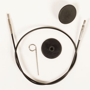 Puede incluir: Un cable negro para agujas de tejer circulares con puntas met&aacute;licas, una peque&ntilde;a herramienta met&aacute;lica y dos topes de goma negros est&aacute;n dispuestos sobre un fondo blanco. El cable es curvo y los topes son redondos.