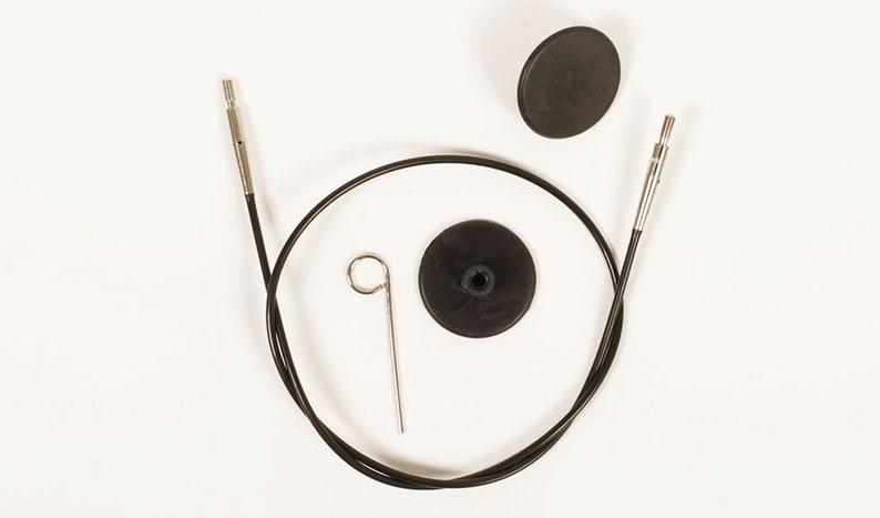 Puede incluir: Un cable negro para agujas de tejer circulares con puntas met&aacute;licas, una peque&ntilde;a herramienta met&aacute;lica y dos topes de goma negros est&aacute;n dispuestos sobre un fondo blanco. El cable es curvo y los topes son redondos.