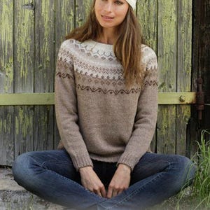 May include: A beige and white long-sleeve knit sweater with a round neckline and a fair isle pattern across the chest and shoulders, paired with a matching knit beanie featuring a pom-pom and blue denim jeans.