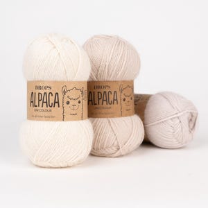 May include: Three skeins of off-white and light beige alpaca yarn. Each skein features a tan paper label with the text DROPS ALPACA UNI COLOUR and a line drawing of an alpaca face. The yarn is wound into a soft, rounded ball shape.