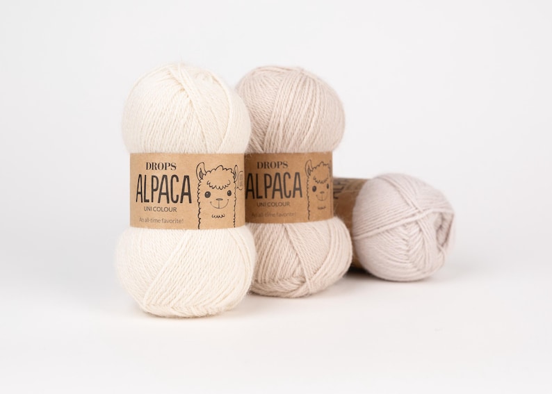 May include: Three skeins of off-white and light beige alpaca yarn. Each skein features a tan paper label with the text DROPS ALPACA UNI COLOUR and a line drawing of an alpaca face. The yarn is wound into a soft, rounded ball shape.