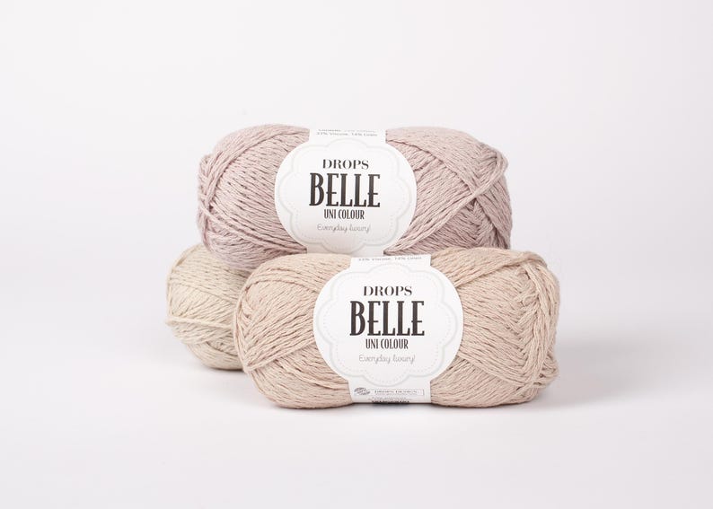 May include: Several skeins of Drops Belle Uni Colour yarn in neutral tones, including beige and light pink. Each skein has a white label with black text. The yarn is arranged on a white background, ideal for knitting and crochet projects.