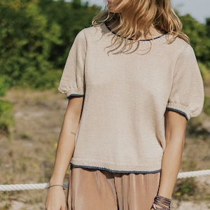 May include: Beige knit top with short, puffed sleeves and navy blue trim. The top is paired with pleated, tan pants. The outfit is worn outdoors on a sunny day. The model is wearing sunglasses and jewelry.