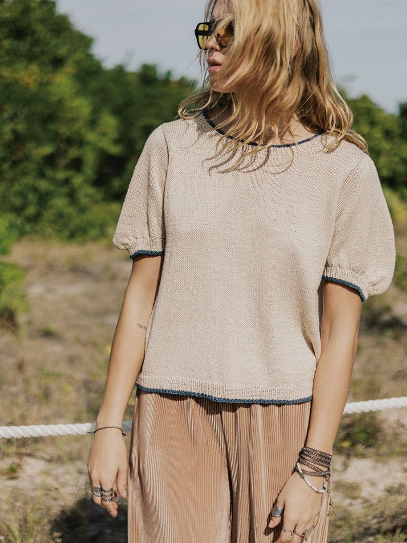 May include: Beige knit top with short, puffed sleeves and navy blue trim. The top is paired with pleated, tan pants. The outfit is worn outdoors on a sunny day. The model is wearing sunglasses and jewelry.