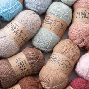 May include: A collection of yarn skeins in various pastel and earth tones, each wrapped in a brown paper label featuring a line drawing of an alpaca and the text DROPS ALPACA. The yarn textures appear soft and fibrous, arranged in a cluster.