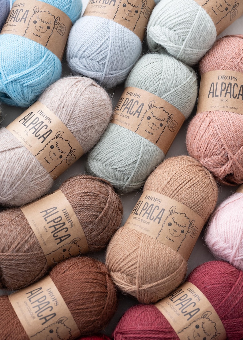 May include: A collection of yarn skeins in various pastel and earth tones, each wrapped in a brown paper label featuring a line drawing of an alpaca and the text DROPS ALPACA. The yarn textures appear soft and fibrous, arranged in a cluster.