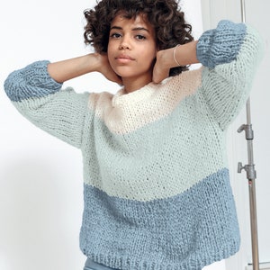 May include: A chunky knit sweater in a color block design. The sweater features a light blue, pale pink, and darker blue color scheme. The sleeves are a darker blue. The sweater is made of a thick, textured yarn.