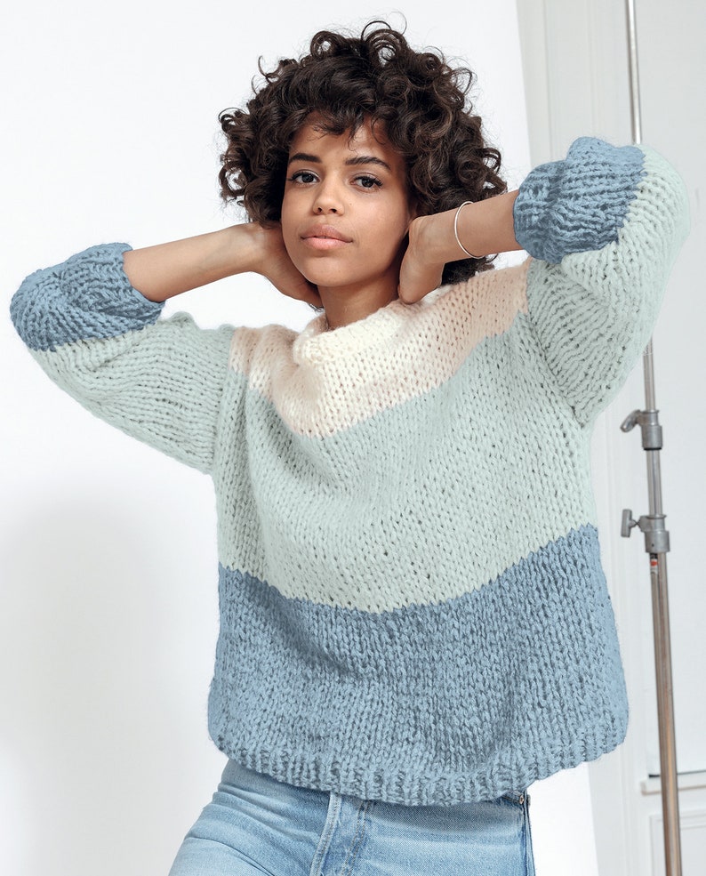May include: A chunky knit sweater in a color block design. The sweater features a light blue, pale pink, and darker blue color scheme. The sleeves are a darker blue. The sweater is made of a thick, textured yarn.