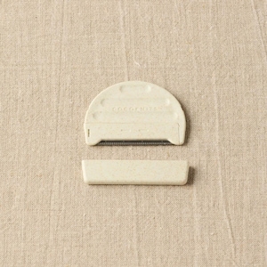 May include: Two-piece, off-white fabric shaver with the text "COCOFRUITS" on the top piece. The top piece is semi-circular with a comb-like blade. The bottom piece is rectangular. The items are on a beige linen surface.