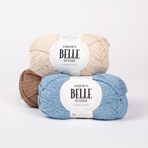 May include: Three skeins of Drops Belle Uni Colour yarn in beige, brown, and blue. Each skein has a label with the brand name and product details. The yarn is arranged in a stack against a white background.