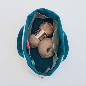 Puede incluir: Un bolso tote de color turquesa y crema, abierto para revelar dos ovillos de lana, un ganchillo rosa y una cinta m&eacute;trica verde. El interior del bolso es turquesa, con un exterior crema y rayas blancas.