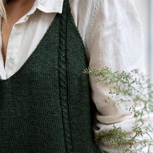May include: A dark green knitted sleeveless top with a cable knit detail on the straps. The top is worn over a white button-down shirt.