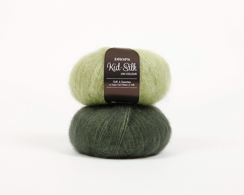 May include: Two balls of Drops Kid-Silk Uni Colour yarn, one light green and one dark green, stacked on top of each other. The top ball has a brown label with white text. The yarn appears soft and fluffy.
