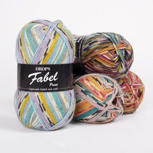 May include: Three balls of Drops Fabel Print yarn. The yarn is a superwash treated sock wool and is multicolored with shades of blue, yellow, orange, pink, green, brown, and black. The label on the yarn reads 'DROPS Fabel Print Superwash treated sock wool.'