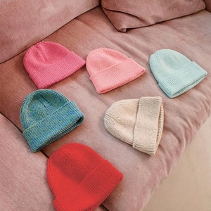 May include: Six knitted beanies in various colours, including pink, blue, red, and cream, are arranged on a pink sofa.