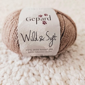 May include: A ball of Gepard Wild & Soft yarn in a light brown hue. The label features the brand name and the text "Wild & Soft". The yarn is composed of 40% wild tussah silk and 60% merino wool. The yarn is resting on a textured, off-white surface.