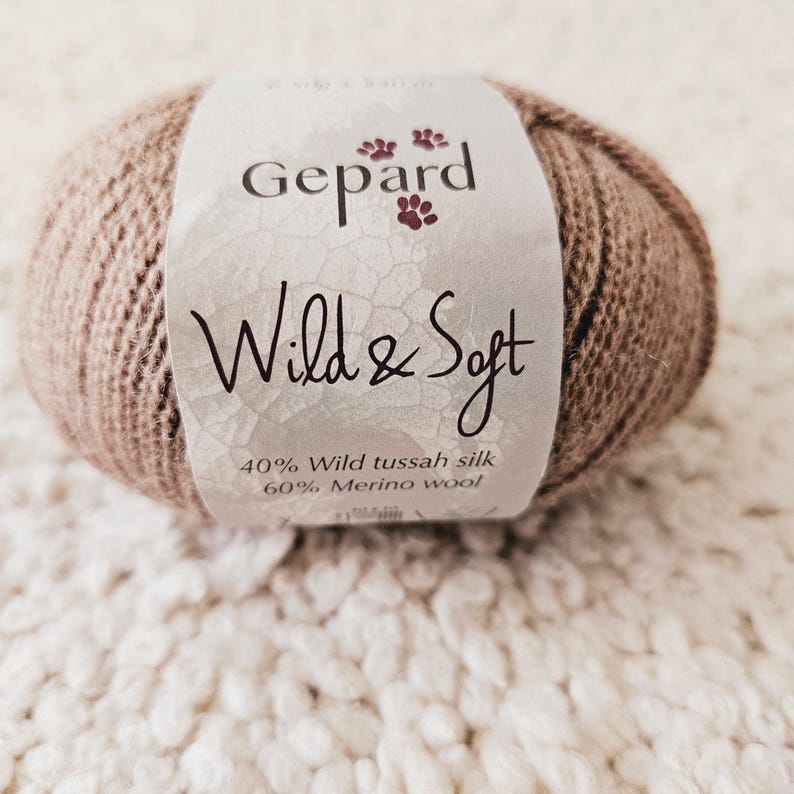 May include: A ball of Gepard Wild & Soft yarn in a light brown hue. The label features the brand name and the text "Wild & Soft". The yarn is composed of 40% wild tussah silk and 60% merino wool. The yarn is resting on a textured, off-white surface.