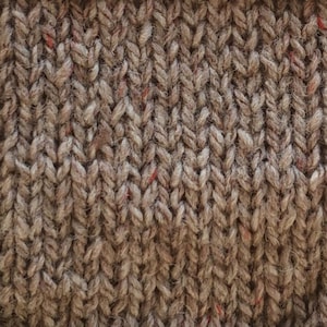 May include: Close-up of a brown knitted fabric with a subtle herringbone pattern. The fabric has a slightly textured surface and a few red flecks throughout.