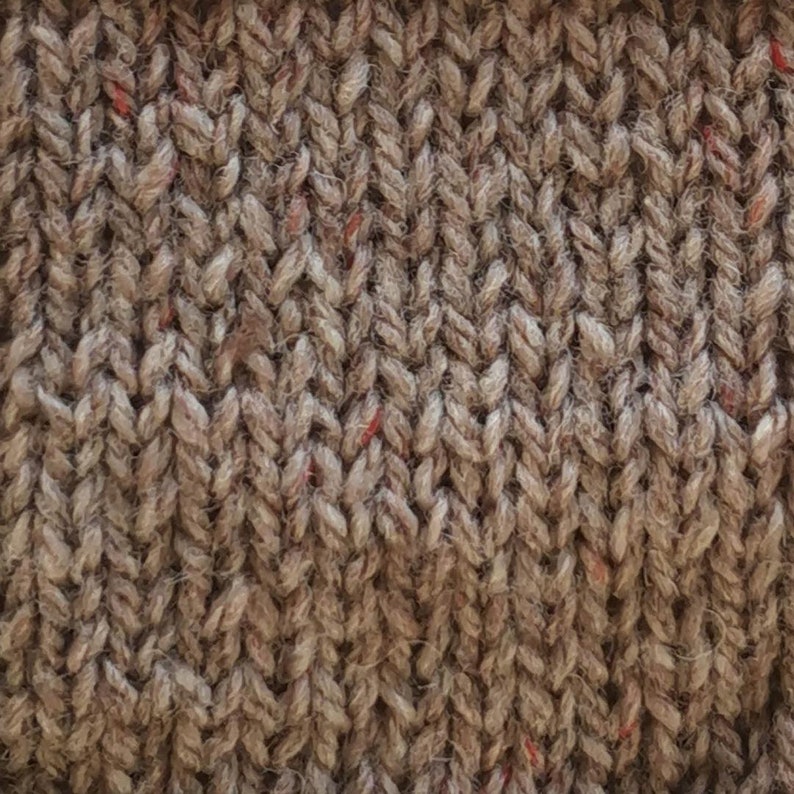 May include: Close-up of a brown knitted fabric with a subtle herringbone pattern. The fabric has a slightly textured surface and a few red flecks throughout.