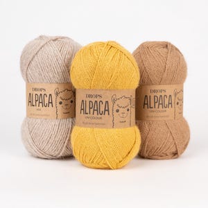 May include: Three skeins of Drops Alpaca yarn in light beige, bright yellow, and medium brown. Each skein features a tan paper label with the text Drops Alpaca, a line drawing of an alpaca face, and the words Mix or Uni Colour printed on the front.