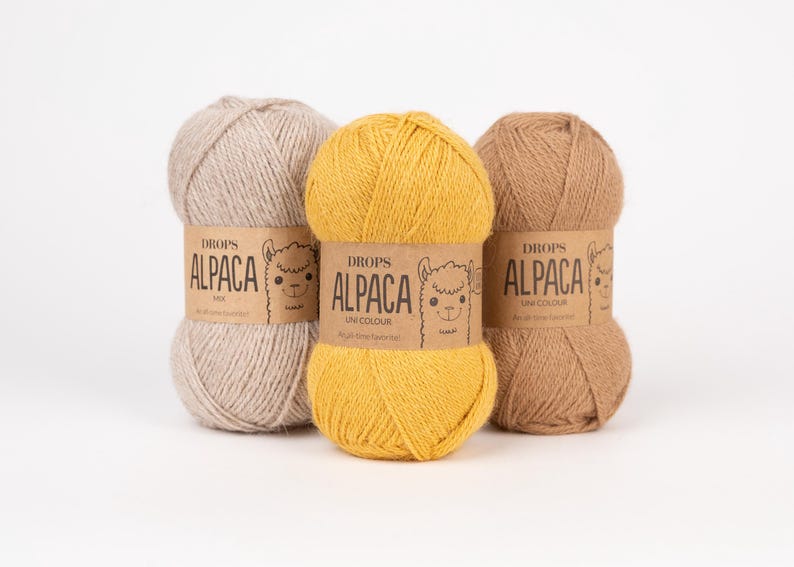 May include: Three skeins of Drops Alpaca yarn in light beige, bright yellow, and medium brown. Each skein features a tan paper label with the text Drops Alpaca, a line drawing of an alpaca face, and the words Mix or Uni Colour printed on the front.