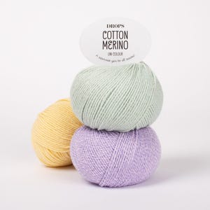Puede incluir: Tres ovillos de lana Drops Cotton Merino en amarillo pastel, verde claro y violeta claro. Una etiqueta ovalada blanca est&aacute; fijada en la parte superior con el texto: DROPS COTTON MERINO UNI COLOUR A superwash yarn for all seasons.