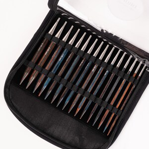 Puede incluir: Un estuche negro con cremallera contiene un juego de agujas de tejer de madera. Las agujas tienen puntas plateadas y mangos de madera en tonos marrones y azules. El estuche tiene una tapa de pl&aacute;stico transparente.