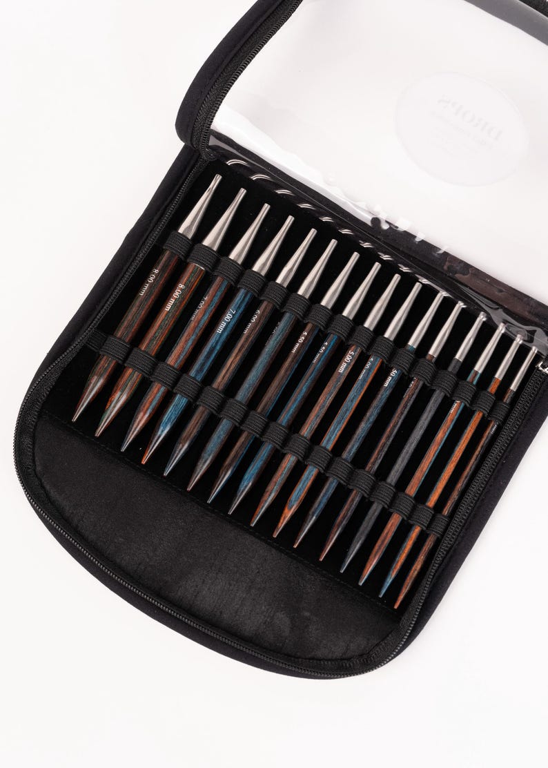 Puede incluir: Un estuche negro con cremallera contiene un juego de agujas de tejer de madera. Las agujas tienen puntas plateadas y mangos de madera en tonos marrones y azules. El estuche tiene una tapa de pl&aacute;stico transparente.