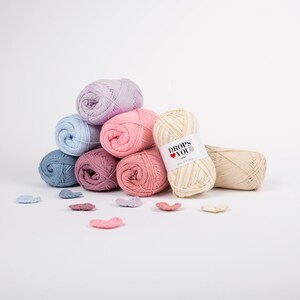 Puede incluir: Una colecci&oacute;n de ovillos de lana coloridos en tonos azules, rosas y morados, dispuestos en forma de pir&aacute;mide. Una etiqueta en uno de los ovillos dice "DROPS YOU". Varios peque&ntilde;os trozos de hilo en forma de coraz&oacute;n est&aacute;n dispersos.