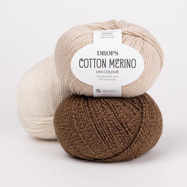 Puede incluir: Tres ovillos de lana Drops Cotton Merino en beige, blanco roto y marr&oacute;n. El ovillo beige superior tiene una etiqueta blanca con el texto: DROPS COTTON MERINO, UNI COLOUR, A superwash yarn for all seasons, Content: 50% Wool, 50% Cotton, Made in EU.