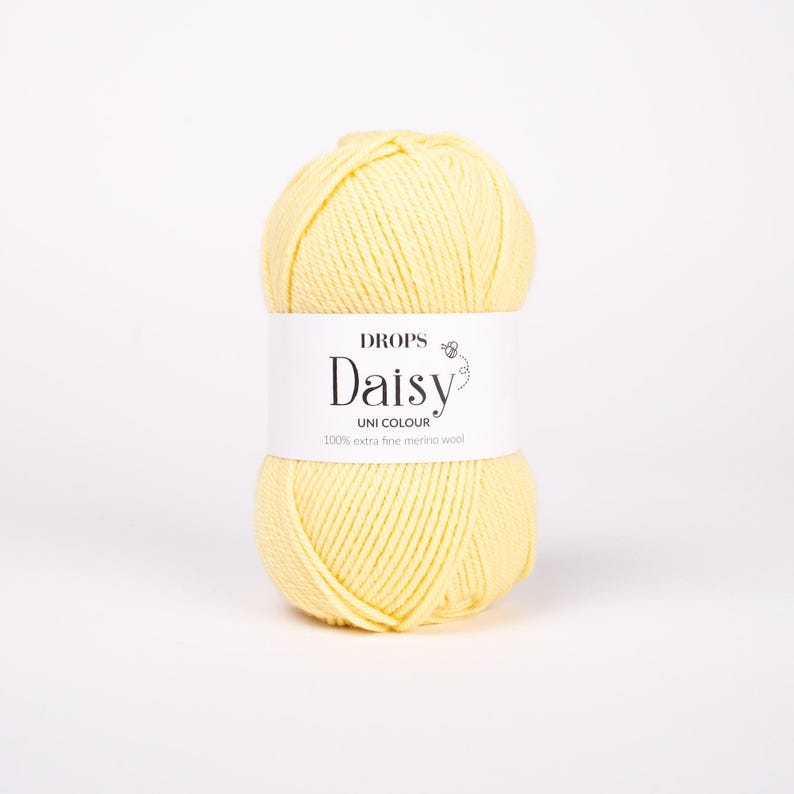 May include: A ball of pale yellow yarn with a white paper label wrapped around the center. The label features the text DROPS Daisy UNI COLOUR 100% extra fine merino wool and a small graphic of a flower. The yarn has a smooth, twisted texture.