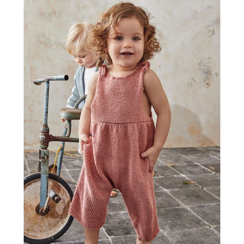 Peut inclure: Un jeune enfant portant une salopette en tricot rose avec des bretelles et une taille fronc&eacute;e. L'enfant est debout devant un tricycle vintage.