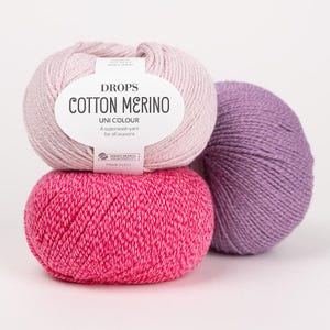 Puede incluir: Tres ovillos de lana Drops Cotton Merino en rosa claro, rosa intenso y morado. El ovillo rosa claro superior tiene una etiqueta blanca con el texto DROPS COTTON MERINO, UNI COLOUR, A superwash yarn for all seasons y Made in EU.