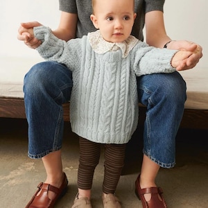 Op de afbeelding: Een baby in een lichtblauwe kabelgebreide trui met een witte kanten kraag, bruin gestreepte leggings en beige schoenen. De baby wordt vastgehouden door een persoon in een grijze trui en een blauwe spijkerbroek. Bruine leren schoenen zijn ook zichtbaar.