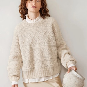 May include: A cream-colored knitted sweater with a diamond pattern. The sweater has long sleeves and a relaxed fit.