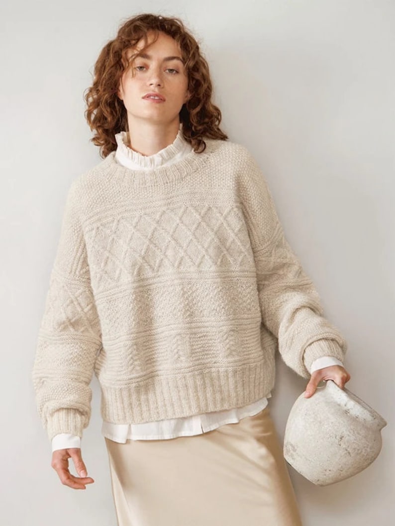 May include: A cream-colored knitted sweater with a diamond pattern. The sweater has long sleeves and a relaxed fit.