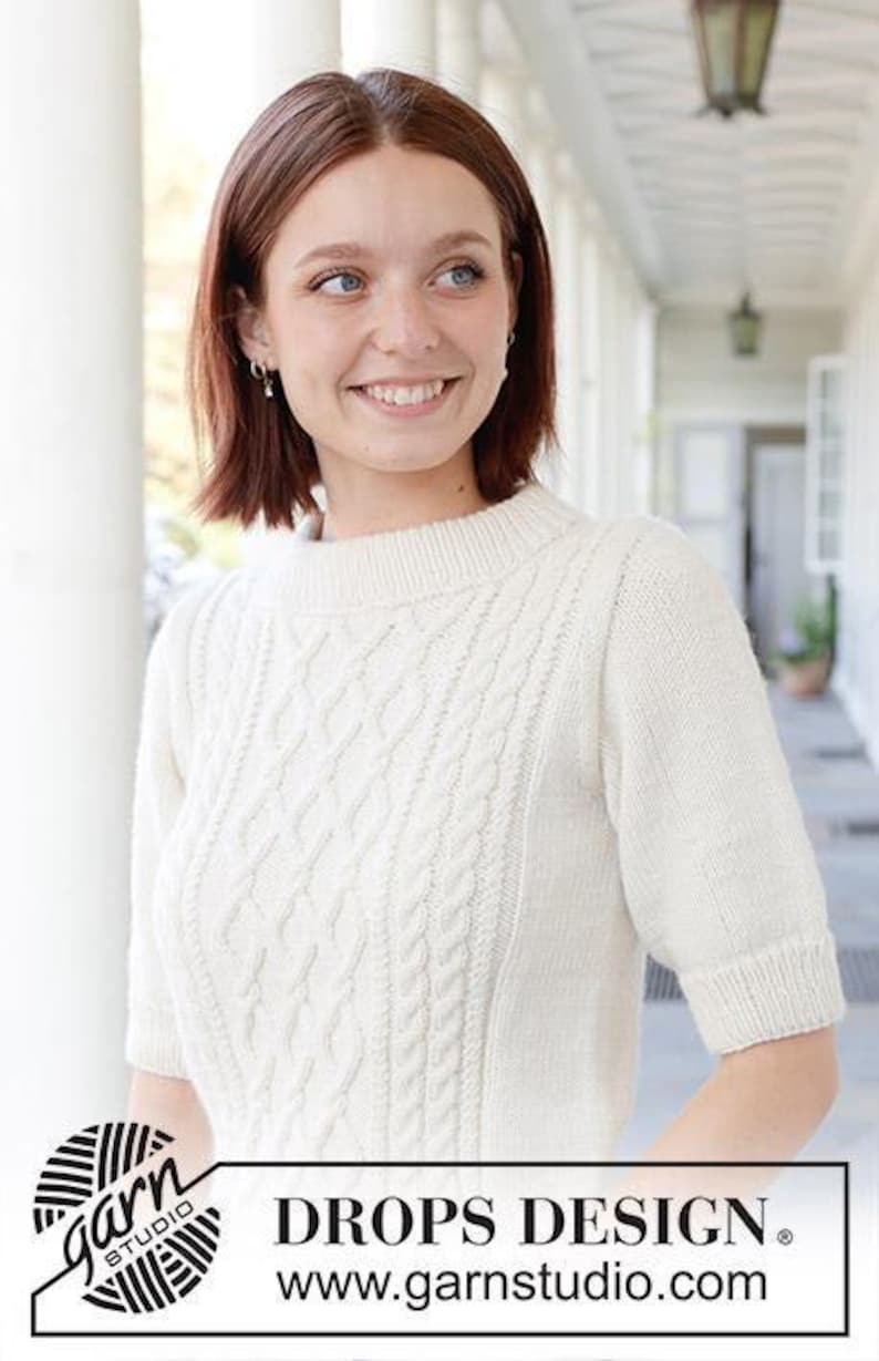 May include: A short-sleeved, off-white knitted sweater featuring a central cable knit pattern on the front, a ribbed crew neckline, and ribbed sleeve cuffs. The image includes the text DROPS DESIGN and the website www.garnstudio.com.