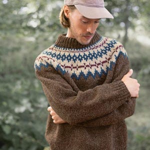 May include: A brown knitted jumper with a geometric pattern in blue, white and red. The jumper has a high neck and long sleeves. The person is wearing a brown baseball cap.