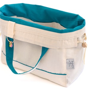 Puede incluir: Bolso tote de color blanco roto con detalles turquesa. El bolso presenta un patr&oacute;n de cuadr&iacute;cula, forro interior turquesa y asas turquesas. Es visible una peque&ntilde;a etiqueta con la palabra "LOMME". Un llavero con abalorios coloridos cuelga de la parte superior.