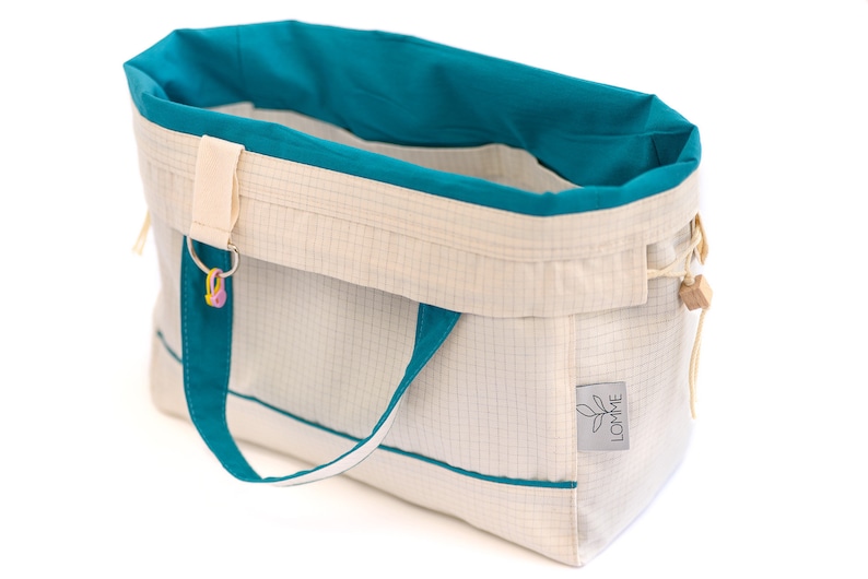 Puede incluir: Bolso tote de color blanco roto con detalles turquesa. El bolso presenta un patr&oacute;n de cuadr&iacute;cula, forro interior turquesa y asas turquesas. Es visible una peque&ntilde;a etiqueta con la palabra "LOMME". Un llavero con abalorios coloridos cuelga de la parte superior.