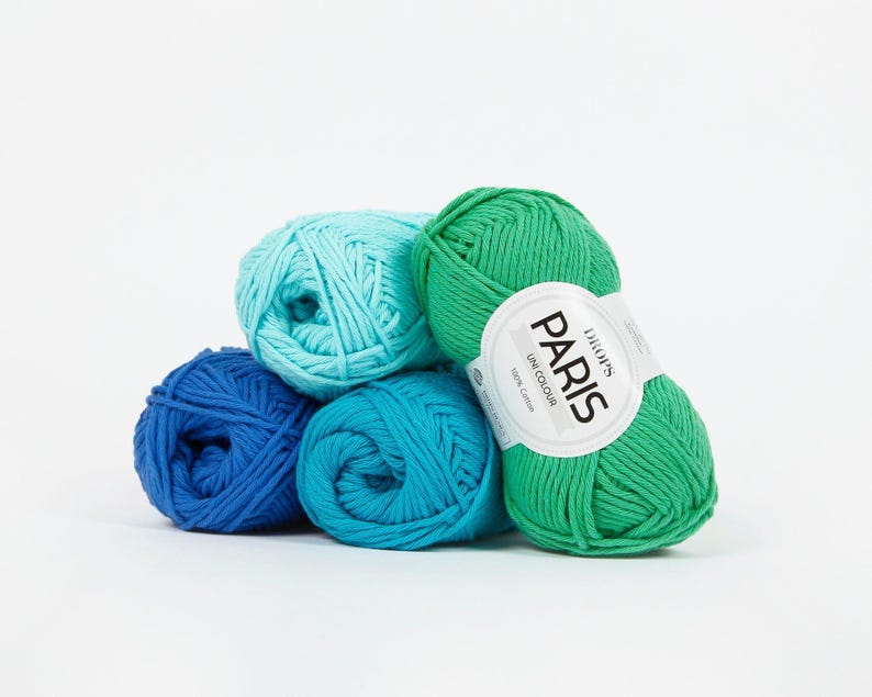 K&ouml;nnte beinhalten: Eine Ansammlung von Garnkn&auml;ueln in verschiedenen Blau- und Gr&uuml;nt&ouml;nen. Ein Kn&auml;uel tr&auml;gt ein Etikett mit der Aufschrift "DROPS PARIS UNI COLOUR 100% Cotton". Die Kn&auml;uel sind vor einem wei&szlig;en Hintergrund gestapelt.