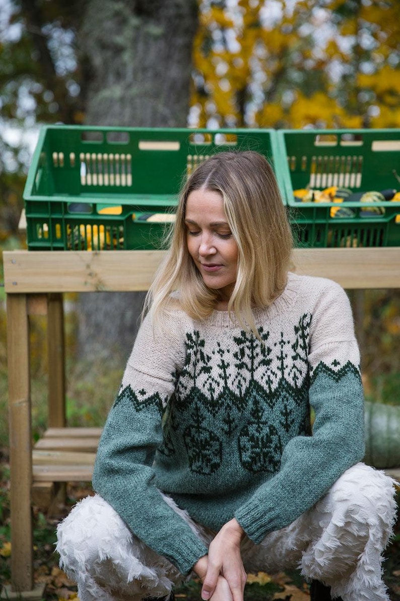 May include: A woman wearing a green and white knitted sweater with a geometric pattern. The sweater has a crew neck and long sleeves. The woman is sitting on a wooden bench in front of a green crate.
