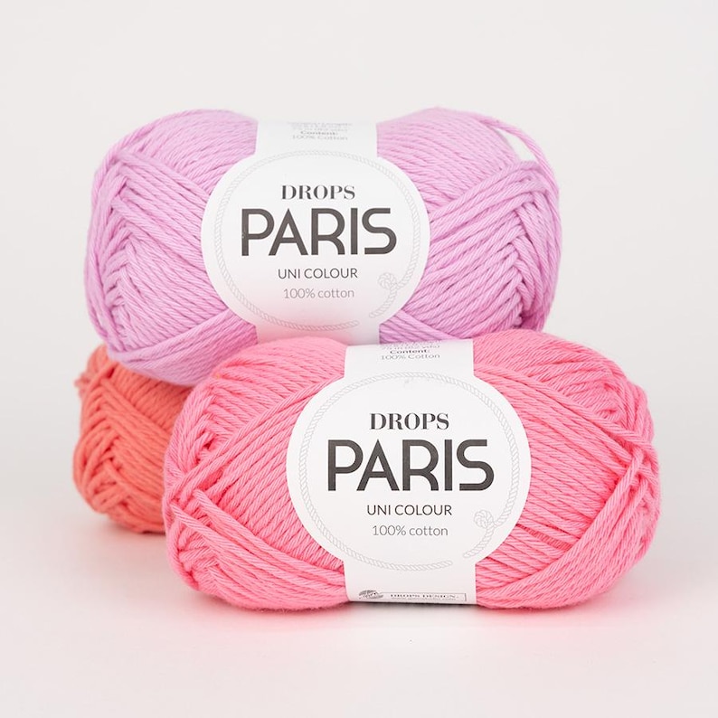 K&ouml;nnte beinhalten: Drei Kn&auml;uel Drops Paris Uni Colour 100% Baumwollgarn in Rosa- und Korallt&ouml;nen. Die Etiketten sind wei&szlig; mit schwarzem Text. Das Garn ist eng gewickelt und zeigt die Textur der Baumwolle. Ideal zum Stricken und H&auml;keln.